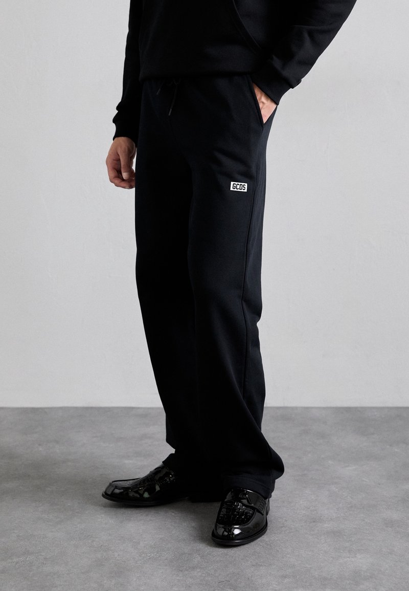 Czarne spodnie dresowe ze ściągaczem w talii i boczną kieszenią, z małą białą metką na górnej części uda. Zestawione z błyszczącymi czarnymi butami.