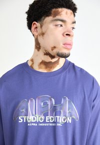 Violet ilgi piedurkņu krekls ar spīdīgu "ALPHA STUDIO EDITION" logo, kas drukāts caurspīdīgās krāsās; gluda kokvilnas tekstūra un klasiskā apkakles dizains.