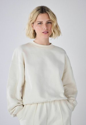 Ung kvinde med blondt hår iført en off-white oversized sweatshirt og matchende bukser, stående mod en ensartet lys baggrund.