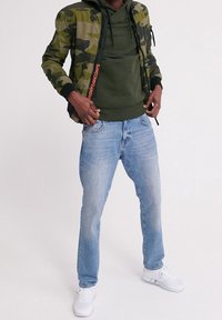 Giacca bomber mimetica, felpa verde con cappuccio, jeans azzurri chiari e sneakers bianche. La giacca presenta dettagli della zip neri e arancioni.