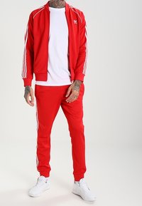 Röd träningsoverall med vita sidostrips. Jackan har en hög krage och dragkedja fram. Byxorna har en avsmalnande passform. Vita sneakers fulländar looken.