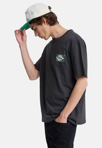 Νέος άνδρας που ρυθμίζει ένα λευκό και πράσινο καπέλο, φορώντας σκούρο γκρι μπλουζάκι με ένα μικρό πράσινο και λευκό σχέδιο στο στήθος, μαύρο παντελόνι, με το χέρι στην τσέπη.