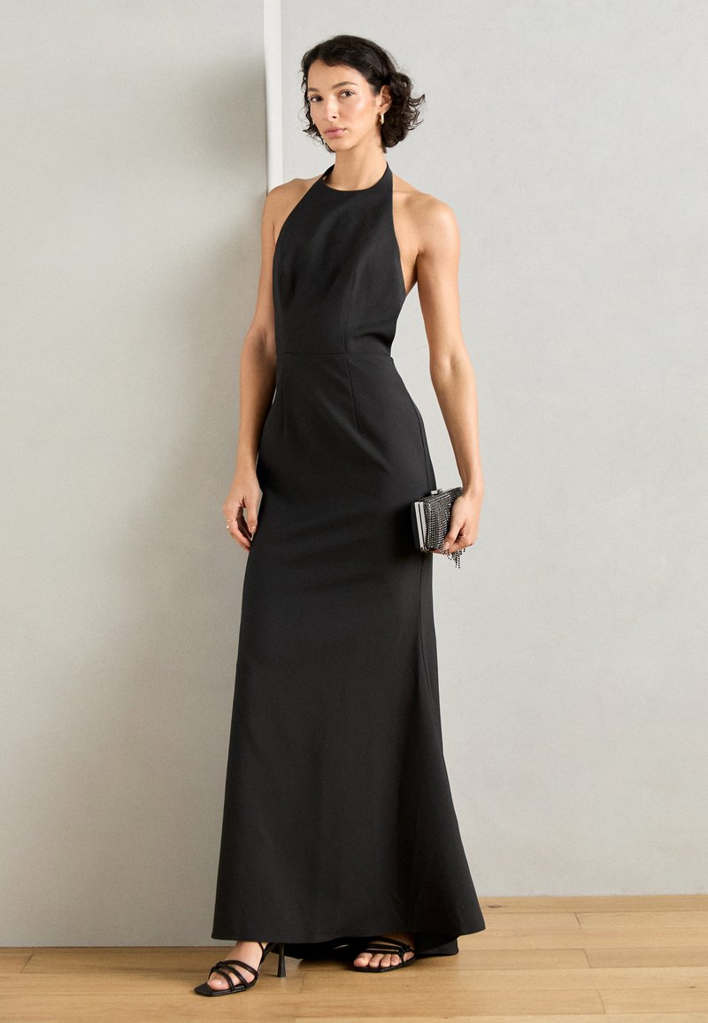 Czarna sukienka na halter z dopasowaną sylwetką i zwiewnym dołem. Dodatki to mała metaliczna kopertówka i czarne sandały na paskach.