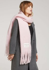 Écharpe en laine rose avec des bords frangés, comportant une étiquette de la marque "Andy & Lucy". Portée lâchement sur un manteau gris.