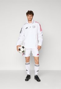 Hvid jakke med sort og rød accent, hvide shorts, sorte fodboldstøvler, holder en sort og hvid fodbold med stjerner, med stribede detaljer.