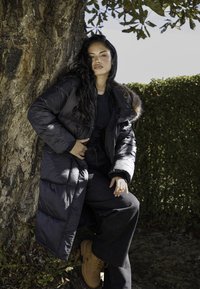 Czarna kurtka puchowa z futrzanym wykończeniem, długa, o błyszczącej fakturze, noszona na czarnej koszulce i luźnych ciemnych spodniach, w połączeniu z jasnobrązowym obuwiem.