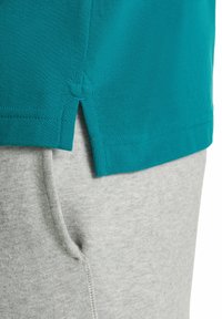 Turkusowy sweter z dzianiny z rozcięciem na dole, noszony na jasnoszarych dresach. Obie garderoby mają teksturowany materiał.