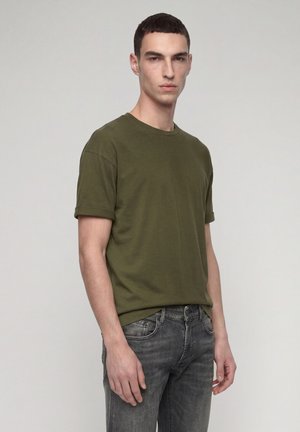 Giovane uomo in piedi davanti a uno sfondo uniforme, indossa una maglietta verde oliva a maniche corte e jeans neri sbiaditi, guarda davanti con espressione neutra.