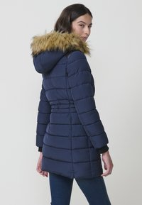 Cappotto piumino blu navy con cappuccio in pelliccia sintetica. Presenta un motivo cucito, maniche lunghe e una vestibilità elegante, progettato per calore e comfort.