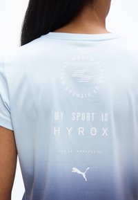 Πίσω μέρος ατόμου που φοράει μπλε αθλητικό μπλουζάκι με διαβάθμιση και το κείμενο «My sport is HYROX» καθώς και το λογότυπο της Puma.