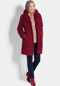 Bordeaux fleece jas met grote kraag, drie knopen en zijzakken, gedragen over een patroontrui en marineblauwe broek, gecombineerd met rode laarzen.