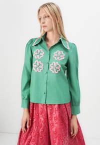 Camisa de algodón verde con aplicaciones florales en volantes rosa, con botones en la parte delantera y cuello grande; presenta mangas abullonadas y una textura suave.