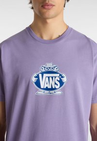 Fialové bavlněné tričko s centrálním modrým logem Vans a kreslenou postavičkou, která drží oválný štítek. Krátké rukávy a design s kulatým výstřihem.