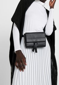 Modelka v černém páskovém batohu přes bílé dlouhé tričko a bílé plisované sukni s černými pruhy, s černým třásňovým šátkem.