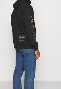 Black hoodie with gold "SWOOSH" text on sleeve and lower back. Features a drawstring hood and ribbed cuffs. Worn with blue jeans.
