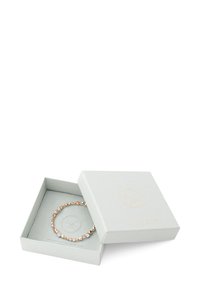 Pulsera en tono dorado con piedras preciosas transparentes, presentada en una caja texturizada de color gris claro con un logo estampado en dorado.