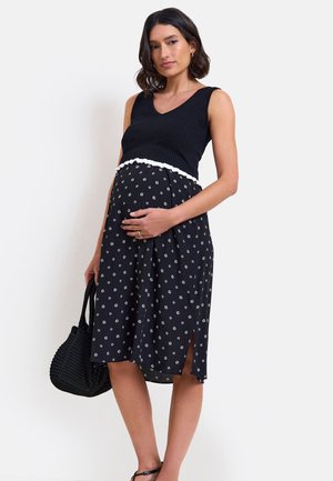 Schwangere Frau in ärmellosem schwarzen Top und gepunktetem Rock, hält mit einer Hand ihren Bauch und mit der anderen eine schwarze Tasche, stehend.