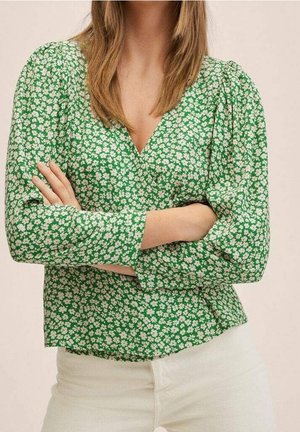 Frau mit verschränkten Armen, die eine grüne Langarmbluse mit weißem Blumenmuster und eine weiße Hose vor beigem Hintergrund trägt.