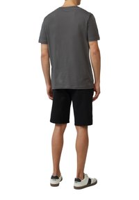 Man die met zijn rug naar de kijker staat, gekleed in een grijs T-shirt met korte mouwen, zwarte shorts en witte sneakers met zwarte accenten.