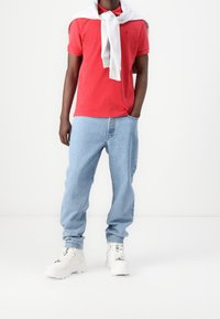 Hombre vestido con una camisa polo roja, jeans azul claros, zapatillas blancas gruesas y un sudadera gris claro atada alrededor de los hombros.