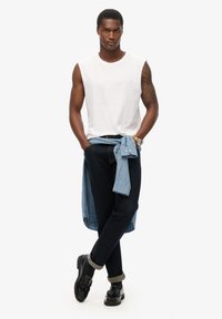 Camiseta blanca sin mangas, jeans de mezclilla oscuros, camisa de mezclilla clara atada a la cintura, mocasines negros con borlas. Fondo liso.