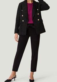 Blazer noir texturé à double boutonnage avec des boutons dorés, associé à un haut à motifs roses et un pantalon noir à taille haute ajusté, complété par des talons noirs.