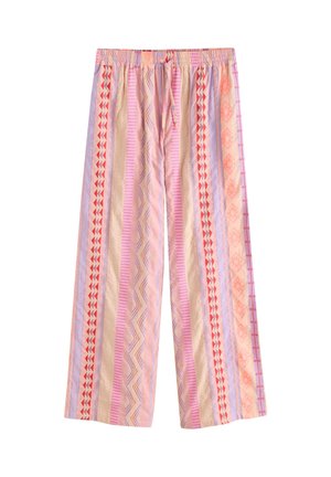 Femme debout contre un mur blanc avec des volets bleus, portant une chemise blanche ouverte, un haut de bikini crème, un pantalon large rose à motifs et des sandales.
