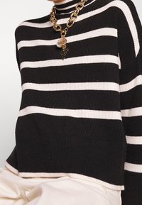 Czarny i biały sweter w poziome paski z prążkowanej tkaniny, zestawiony z masywnym złotym naszyjnikiem łańcuchowym z zawieszkami w kształcie monety, perły i krzyża.