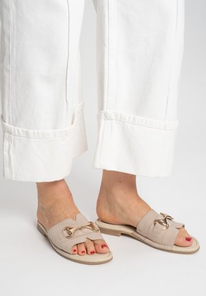 Füße in beigen, offenen Pantolette mit goldenen Akzenten, kombiniert mit weißen, weit geschnittenen Hosen, die über den Knöcheln enden.