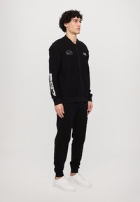 Black bomber jacket with a zip, featuring embroidered logos and white graphics on the sleeves, paired with matching black pants and white sneakers.