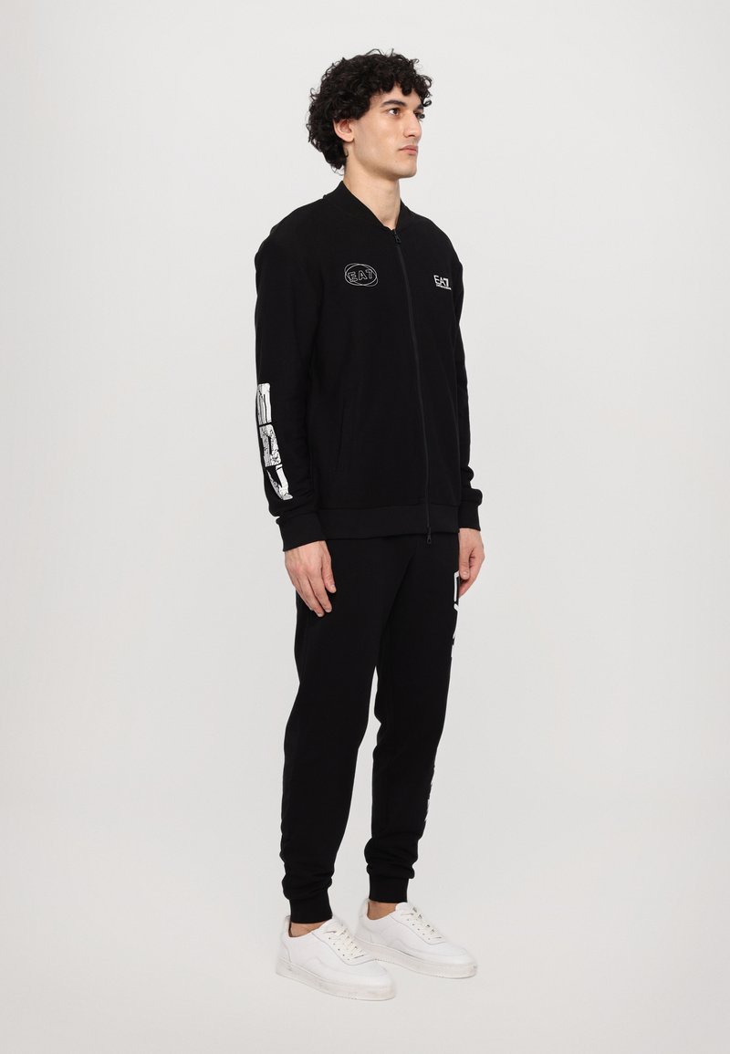 Chaqueta bomber negra con cremallera, con logotipos bordados y gráficos blancos en las mangas, combinada con pantalones negros a juego y zapatillas blancas.