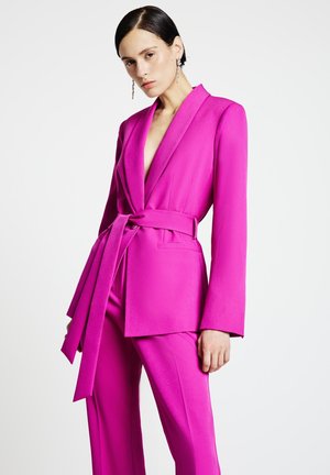 Donna con blazer rosa acceso con cintura abbinato a pantaloni coordinati, capelli tirati indietro e lunghi orecchini d'argento, su sfondo bianco uniforme.