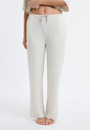 Mujer con pantalones de chándal gris claro con cordón, de pie descalza, levantando una mano para mostrar la barriga.