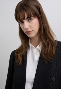 Sort kavaljetjakke med to knapper over en hvid skjorte med krave. Kvinden har langt brunt hår og blå øjne og ser direkte ind i kameraet.