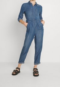 Mono de mezclilla con mangas largas, dos bolsillos en el pecho, cintura elástica y piernas ajustadas. Combinado con sandalias negras de tiras. Color azul claro.