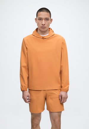 Muž v oranžovej kapucni a zodpovedajúcich oranžových kraťasoch stojaci pred jednoliatym svetlým pozadím, otočený smerom dopredu.