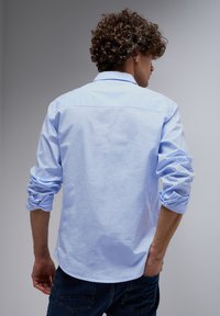 Light blue long-sleeve shirt made of cotton, featuring rolled-up sleeves, a pointed collar, and a curved hem, paired with dark jeans.