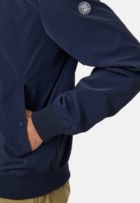 Marineblå let bomberjakke med teksturerede strikkede manchetter, sidelommer og et rundt logo på ærmet. Glat materiale med en tætsiddende design.
