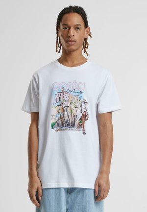 Hvid bomulds-t-shirt med et farverigt frontmotiv, der viser en kystscene med stiliserede bygninger, en klippe og en figur.
