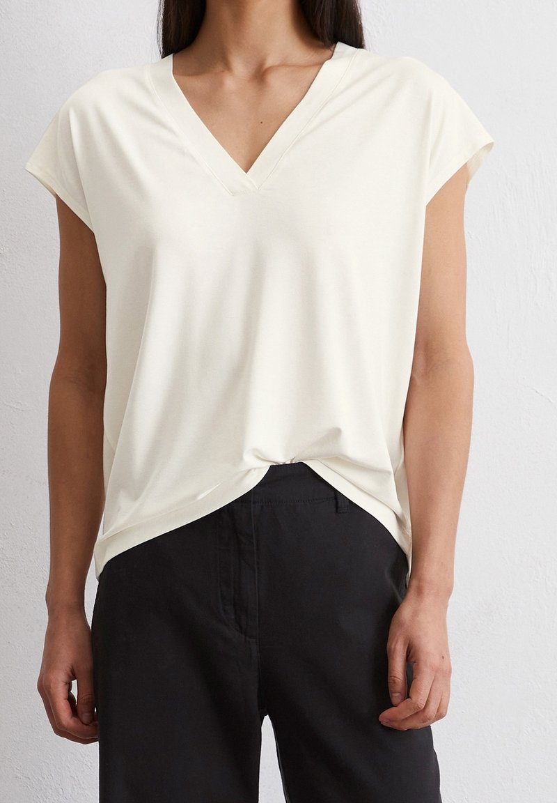 Femme portant un haut blanc à manches courtes en V, légèrement rentré dans un pantalon noir taille haute, devant un mur blanc uni.