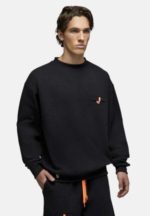 Jeune homme portant un sweat-shirt noir oversize avec une petite poche et un détail orange, associé à un pantalon noir avec des cordons orange vif.
