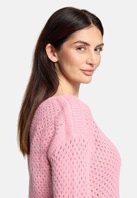 Femme aux longs cheveux foncés portant un pull en tricot rose, regardant par-dessus son épaule avec un léger sourire sur un fond blanc uni.