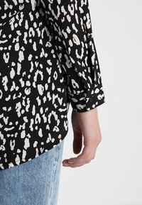 Košile s leopardím vzorem v černobílé barvě s knoflíkovým manžetovým rukávem, hladkou texturou a uvolněným střihem. Viditelná ruka držící okraj košile.