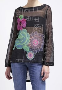 Blouse noire transparente avec une broderie florale vibrante en rose, vert et violet, à manches longues et un ourlet asymétrique.
