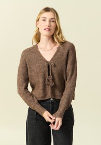 Maglione corto in maglia marrone con scollo a V, chiusura con nodo davanti e maniche lunghe. Tessuto strutturato e polsini a coste.