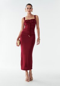 Bordový, priliehavý maxi šaty s volánovým výstrihom a nastaviteľným šnúrkovým pásom. Nosené so zlatými doplnkami a sandálmi na remienky.