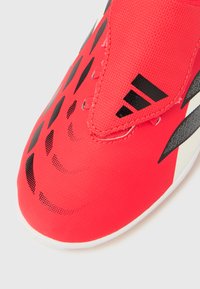 Κοντινό πλάνο από το μπροστινό μέρος ενός κόκκινου αθλητικού παπουτσιού με μαύρα γεωμετρικά σχέδια και λουράκι με αγκράφα, που διαθέτει ένα μαύρο τριγωνικό λογότυπο.