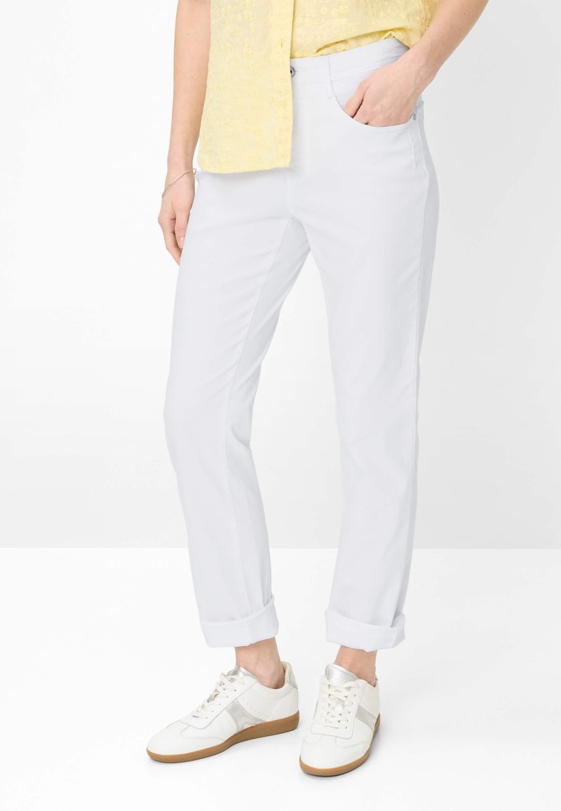 Personne portant un pantalon blanc retroussé, des baskets blanches avec des accents beige, et une chemise jaune à manches courtes rentrée d'un côté.