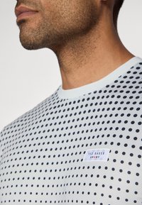 Korte mouwen witte t-shirt met marineblauwe stippen en een ronde halslijn. Heeft een gestikt label met de tekst "TED BAKER SPORT" op de borst.