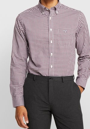 Man wearing long-sleeve maroon and white checkered button-up shirt tucked into dark gray pants, hand in pocket.